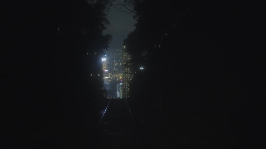 A camera view slowly moves down dark funicular tracks framed by silhouetted trees toward a brightly lit towering city skyline at night
