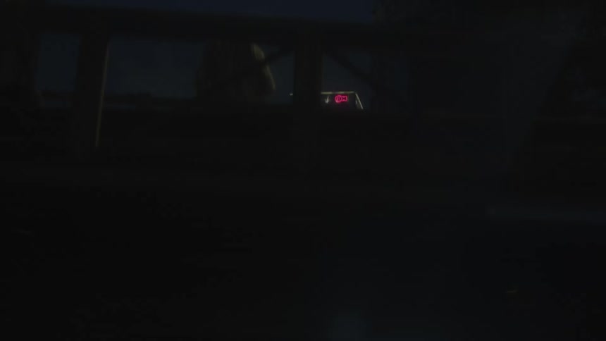 A panning view from inside a vehicle at night observing a tall brightly illuminated rectangular structure and nearby buildings with lit signs framed by dark foreground elements