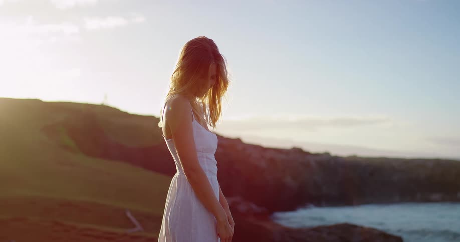 Woman in white dress on coastal cliff at sunset overlooking the sea