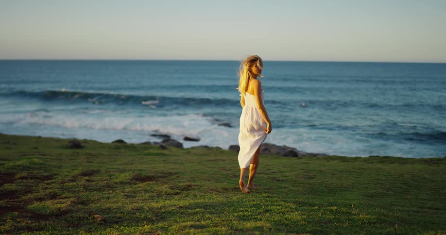 Woman walks barefoot on the grass toward the sea at sunset