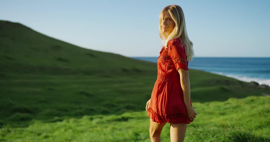 Woman in a red dress walks through a green field overlooking the sea