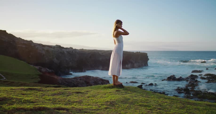 Woman stands on the grass watching the rough sea at sunset
