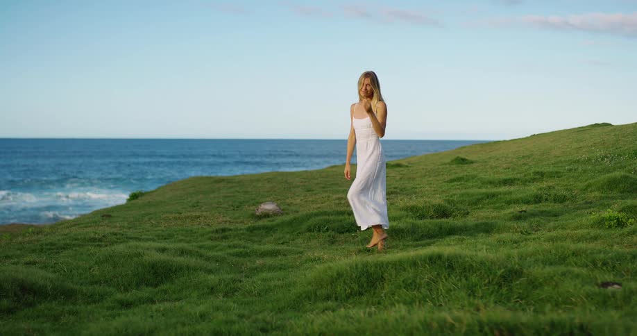 Woman in a white dress walks barefoot on the grass toward the sea