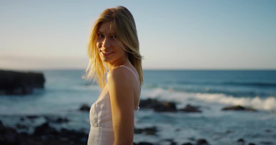 Woman in a white dress looks gently back by the seaside at sunset