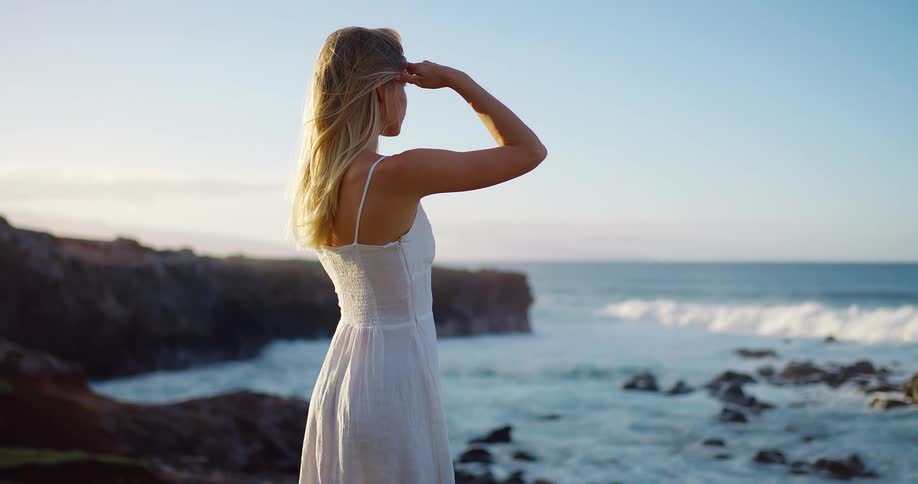 Woman stands on a rocky coastline overlooking ocean waves under a bright sky