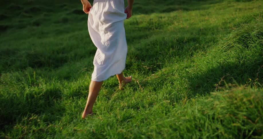 Woman walks barefoot on grassy hill at sunset wearing a white dress and bathed in soft light