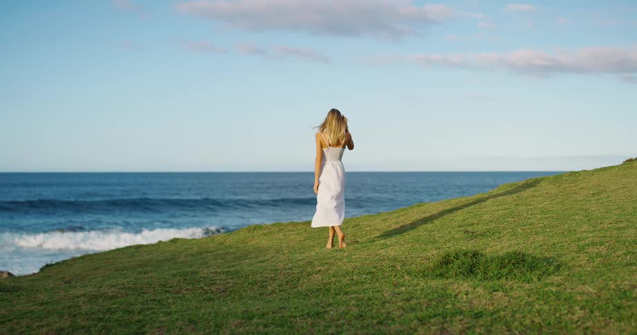Woman walks barefoot on the grassy slope toward the sea under a blue sky