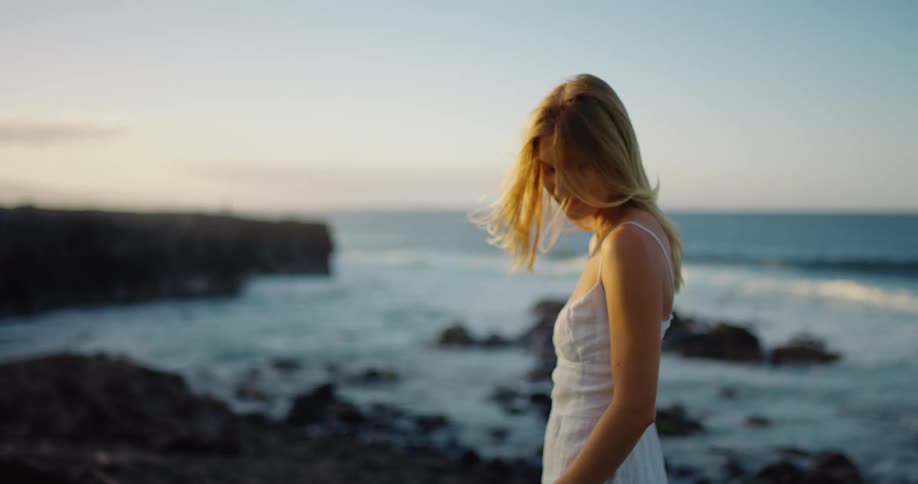 Woman in a white dress touches her hair while enjoying the seaside view at sunset