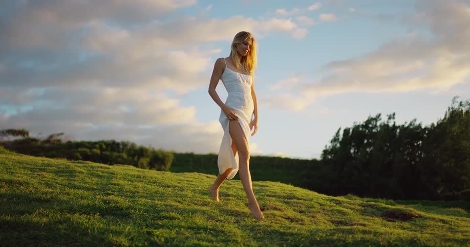 Woman in a white dress walks barefoot across a hill lit by the golden light of sunset