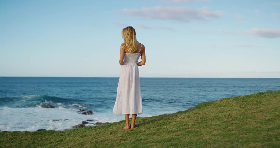 Woman gazes at the sea horizon from a grassy slope under a blue sky