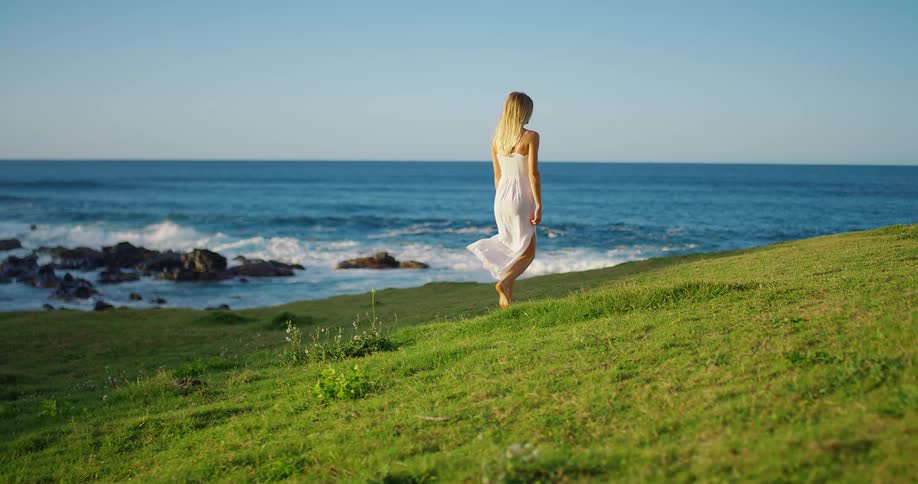 Woman in a white dress walks barefoot on a grassy seaside under the blue sky