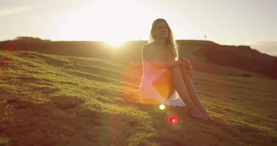 Happy woman sitting on grassy hillside as sun shines in the distance