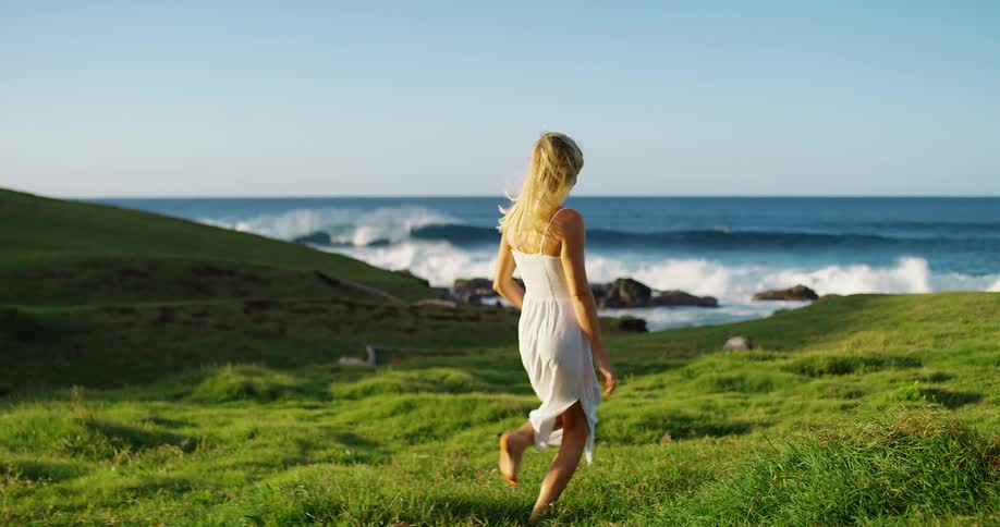 Woman walks barefoot across the grass toward the sea under a clear sky