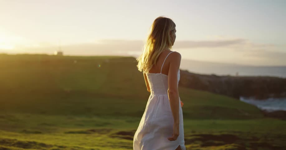 A woman in a white dress runs in slow motion across a grassy cliffside at sunset