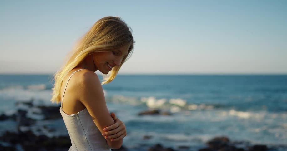 A beautiful blonde woman wearing a white sundress stands on the rocky shoreline turning and smiling at the camera with the blue ocean waves crashing behind her