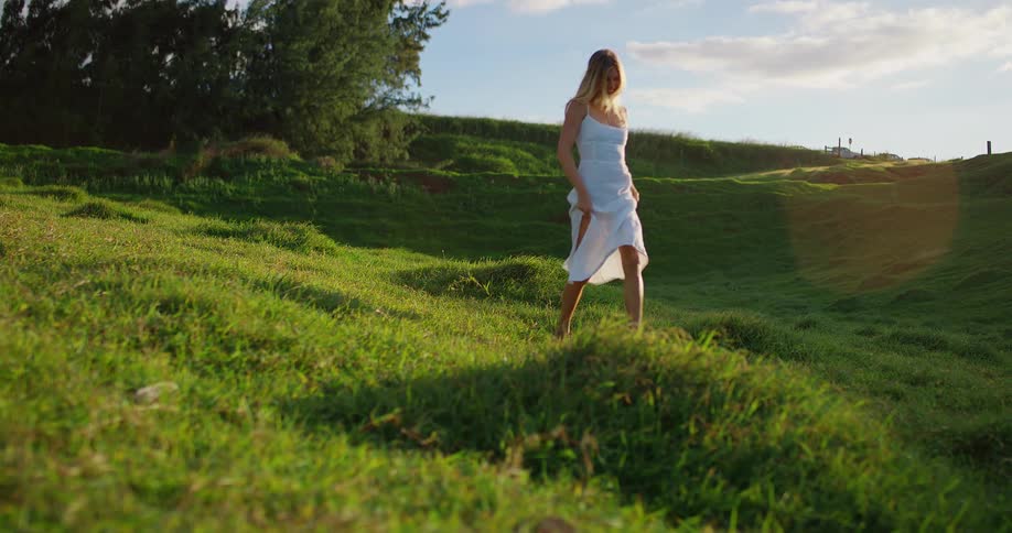 Woman in a white dress walks across a green hill under the soft afternoon light