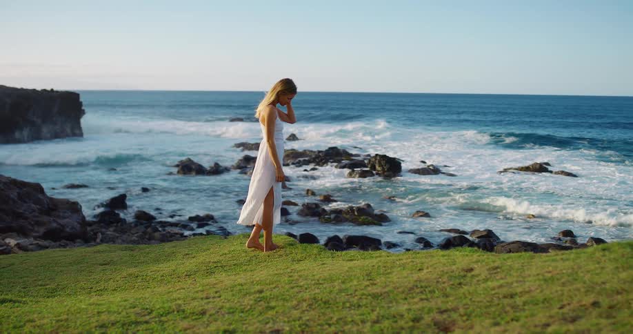 Woman stands barefoot on the grassy slope contemplating the ocean waves