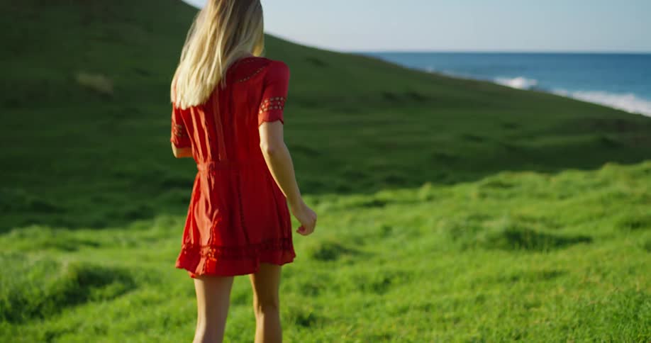 Woman in a red dress walks smiling through a green field by the sea under a clear sky
