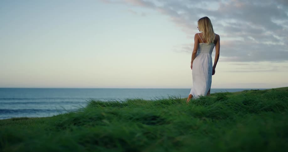 Woman walks on the grass toward the sea at sunset