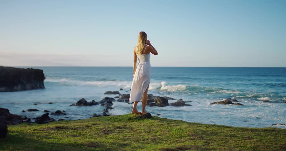 Woman gazes at the horizon by the sea on a grassy cliff under a clear sky