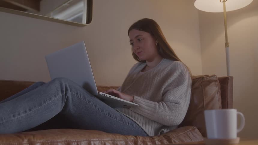 Woman sits comfortably on leather couch using laptop in cozy setting with soft lighting and mug on the table
