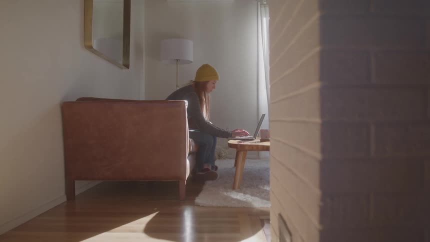 Woman wearing a yellow beanie uses a laptop while sitting on the sofa in a sunlit room
