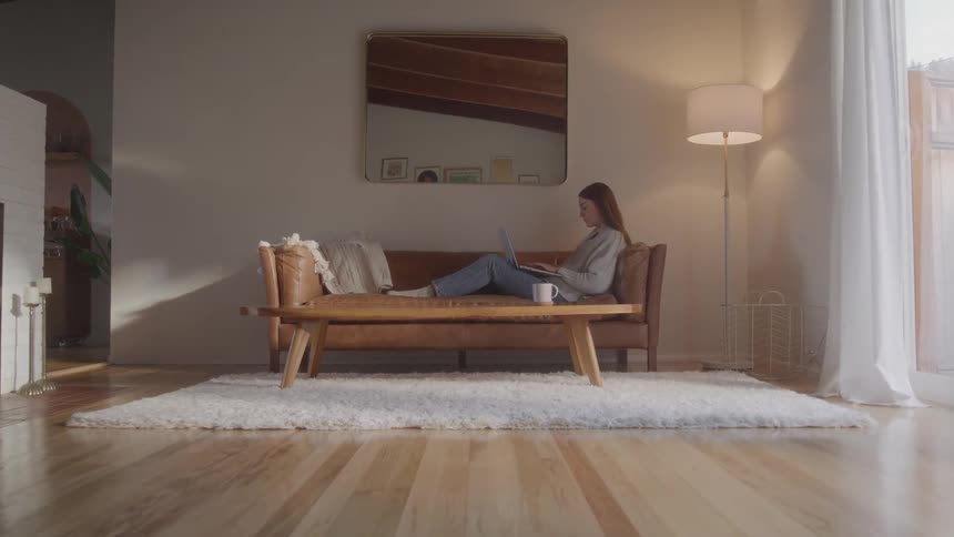 Woman lying on the sofa works on a laptop in a room lit by natural light