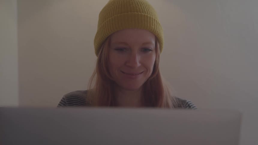 A young woman with long brown hair types on a computer while wearing a yellow beanie
