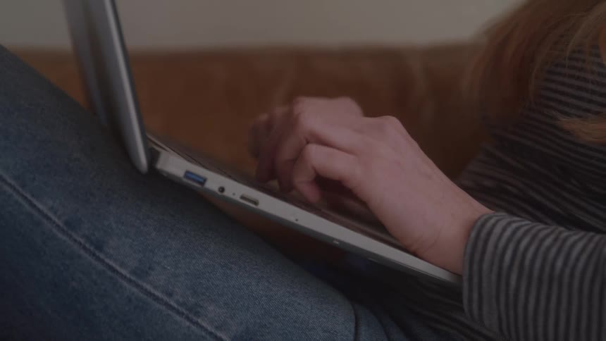 A female sitting on a couch is typing on a laptop that is on her lap