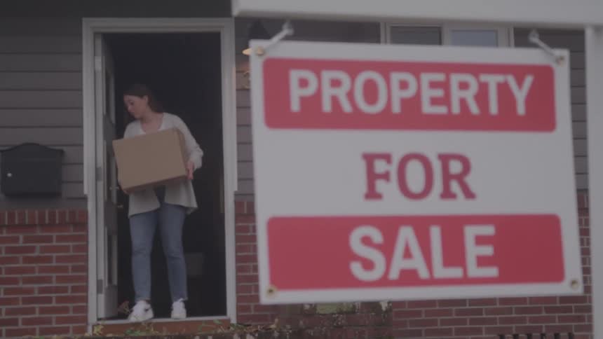 A young woman carries moving boxes out of a building with a property for sale sign outside at midday