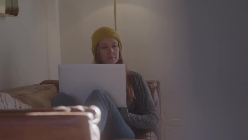 Woman sitting in a loveseat pauses to think before continuing to type on her laptop