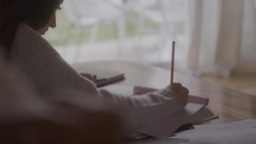 A female is writing with a pencil on paper at a table in a room