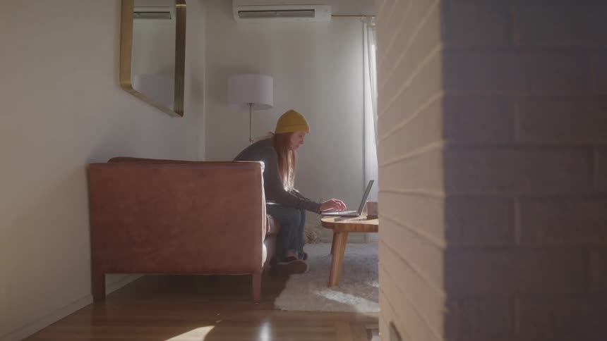 Woman wearing a yellow beanie works on a laptop while sitting on a sunlit sofa