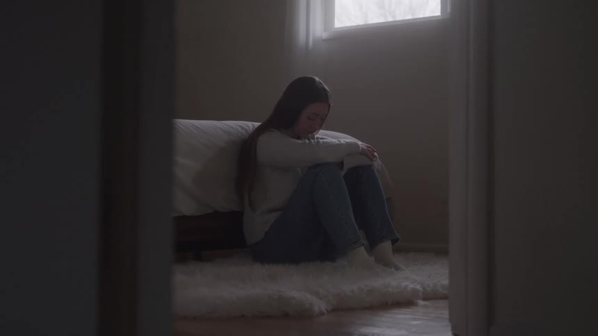 A young woman sits on a shaggy white rug beside a low bed knees hugged to chest head bowed appearing sad and withdrawn in dim window light