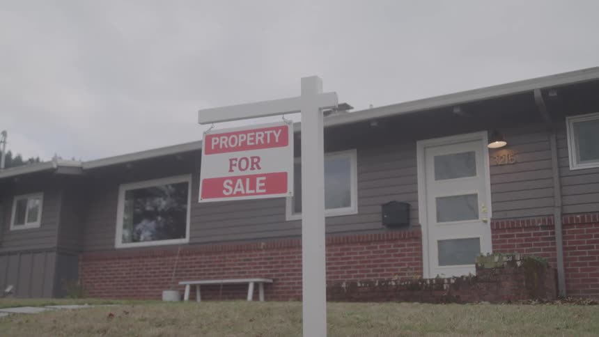 A single story house stands with a for sale sign in its front yard