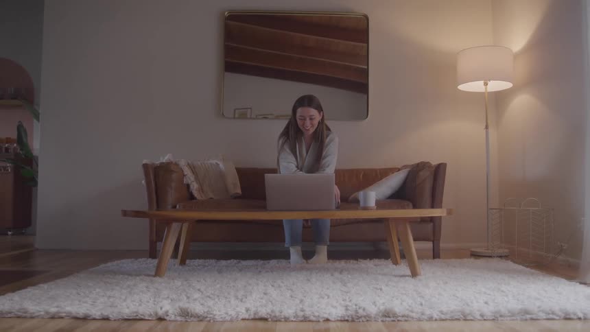 A young woman works on her laptop while sitting on the couch in her living room at midday