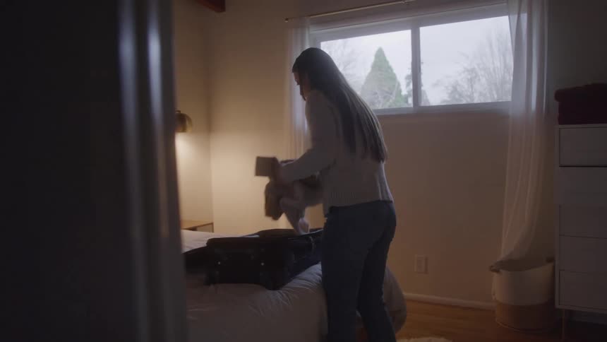 A woman packs clothes into a suitcase on a bed in a sunlit bedroom