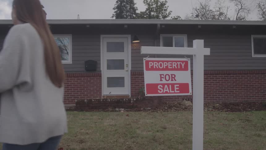 Woman carries a box while passing a property for sale sign in front of a house