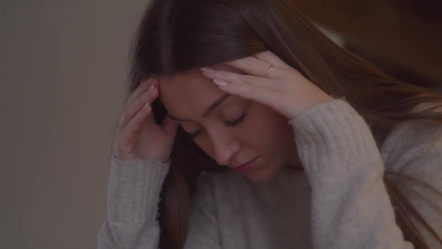 A young woman with long brown hair holds her head in her hands looking down with visible stress and fatigue