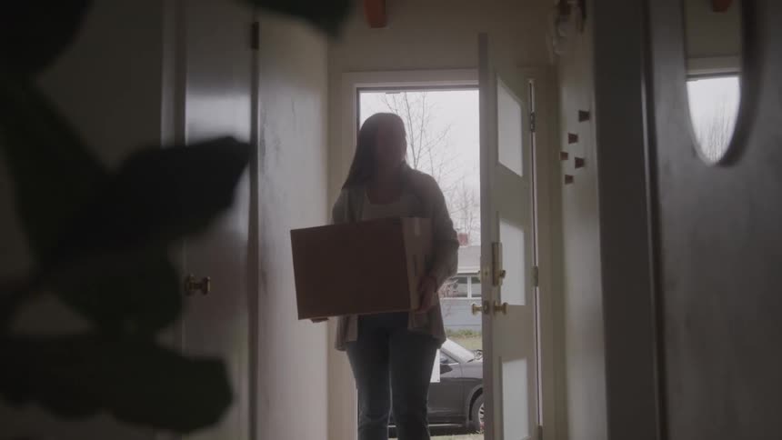 Woman enters through the door carrying a box in a space lit by outdoor light
