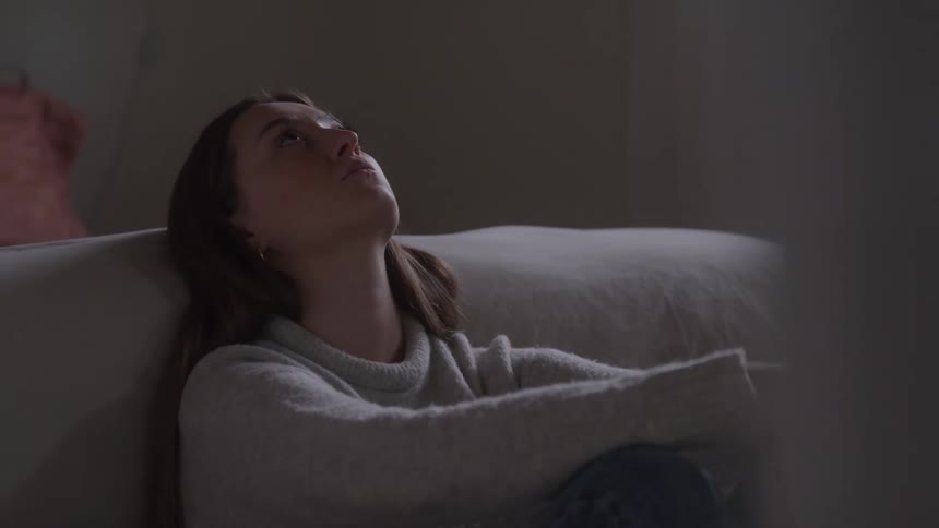 A young woman sits slouched against a couch with her arms wrapped around her knees appearing distant and troubled