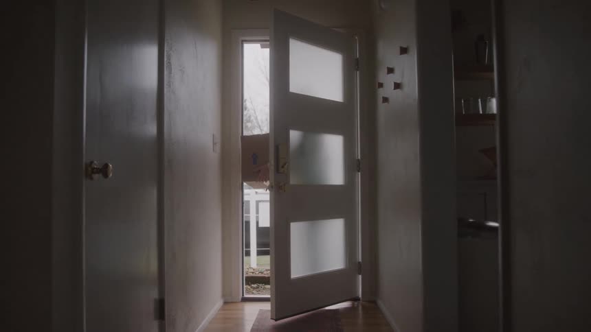 A young woman carries a shipping box through the front door into the house at midday