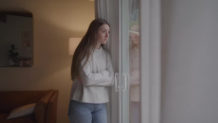 A young woman stands by a window with arms crossed gazing outside appearing pensive and slightly worried