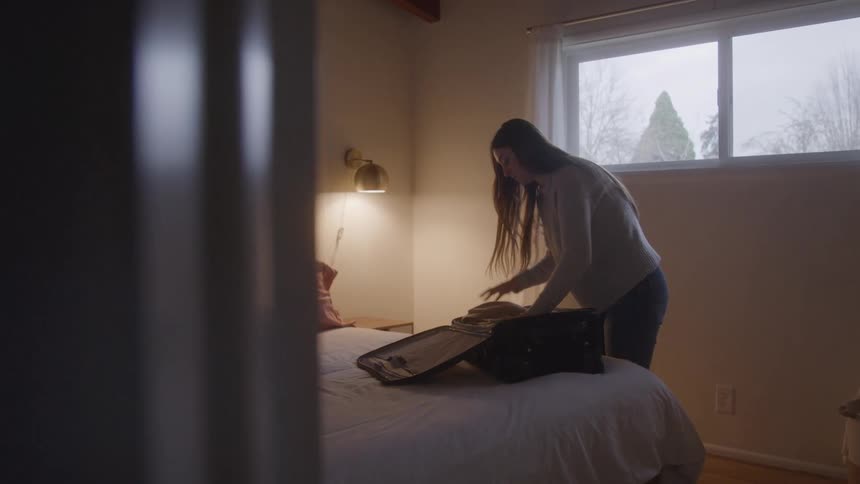 A woman packs an open suitcase in a dimly lit bedroom
