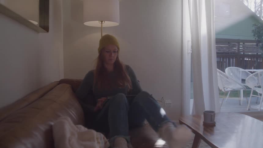 Woman in yellow hat sits on brown sofa and uses laptop