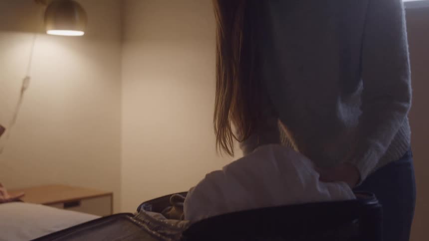 A woman hurriedly packs clothes into an open suitcase on the bed her movements suggesting stress and urgency