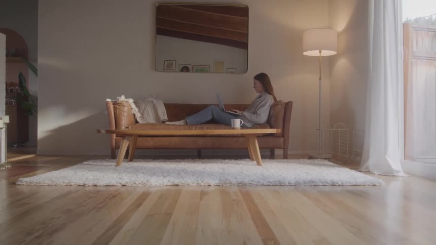 A woman reclines on a tan midcentury sofa with a laptop in a sunlit living room appearing calm and focused