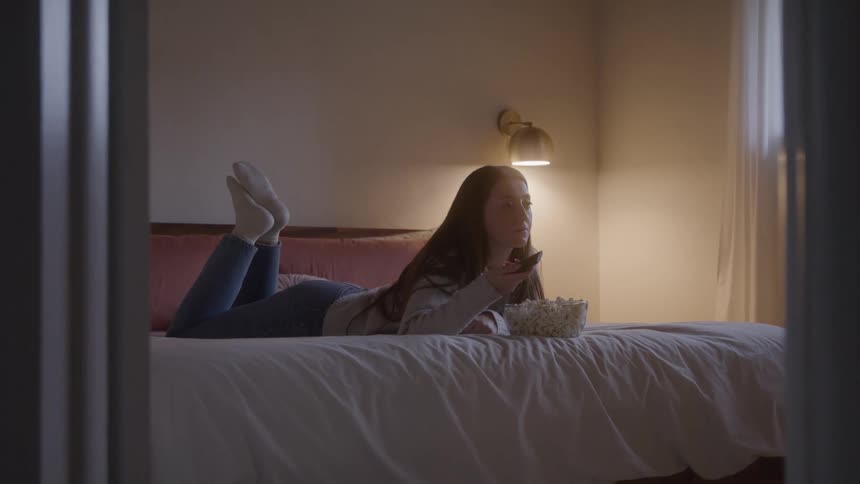 The woman lays in bed eating popcorn and watching television