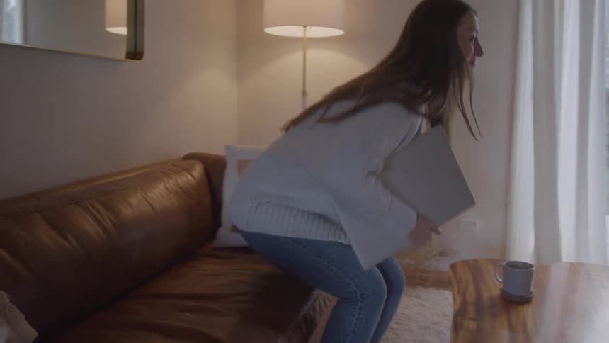 A female walks from a kitchen to the living room carrying a laptop and mug she then reclines on a couch and places laptop in her lap