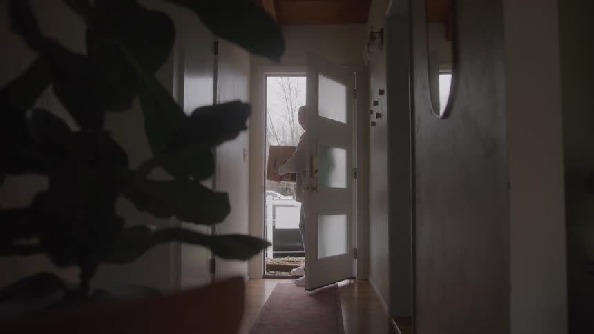 A female carrying a large box walks through a house door into the hallway